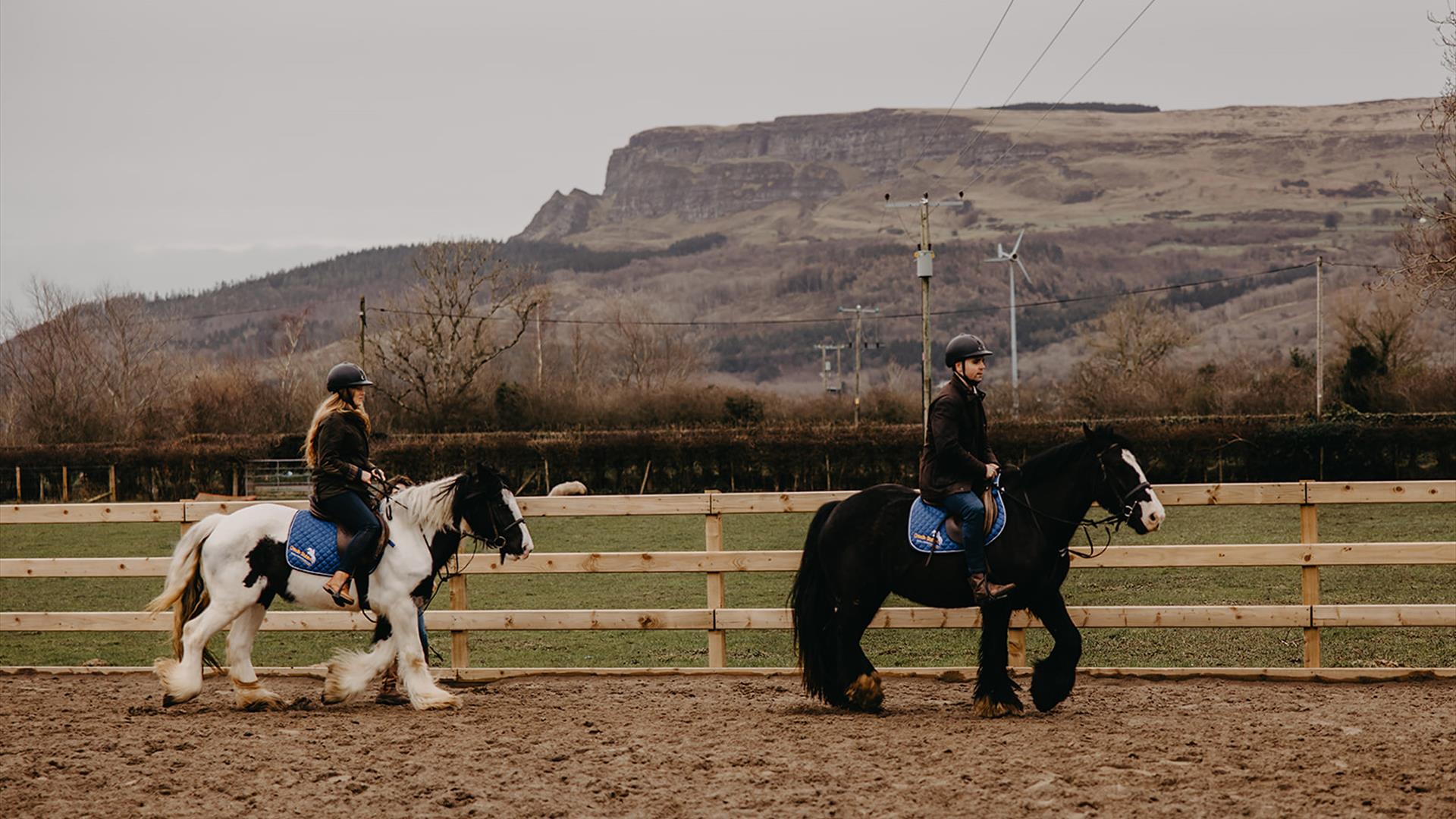 A man on a black horse and a woman on a white horse trotting around an arena with Binevenagh mountain in the background