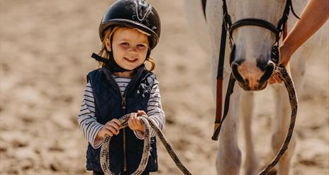 A little girl with a helmet on holding the reins of a white pony