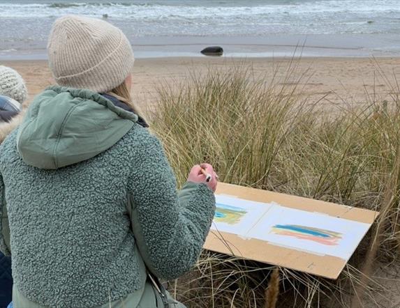 Image of a woman standing in the sand dunes looking out towards the beach, holding a pencil and painting on a colouring board
