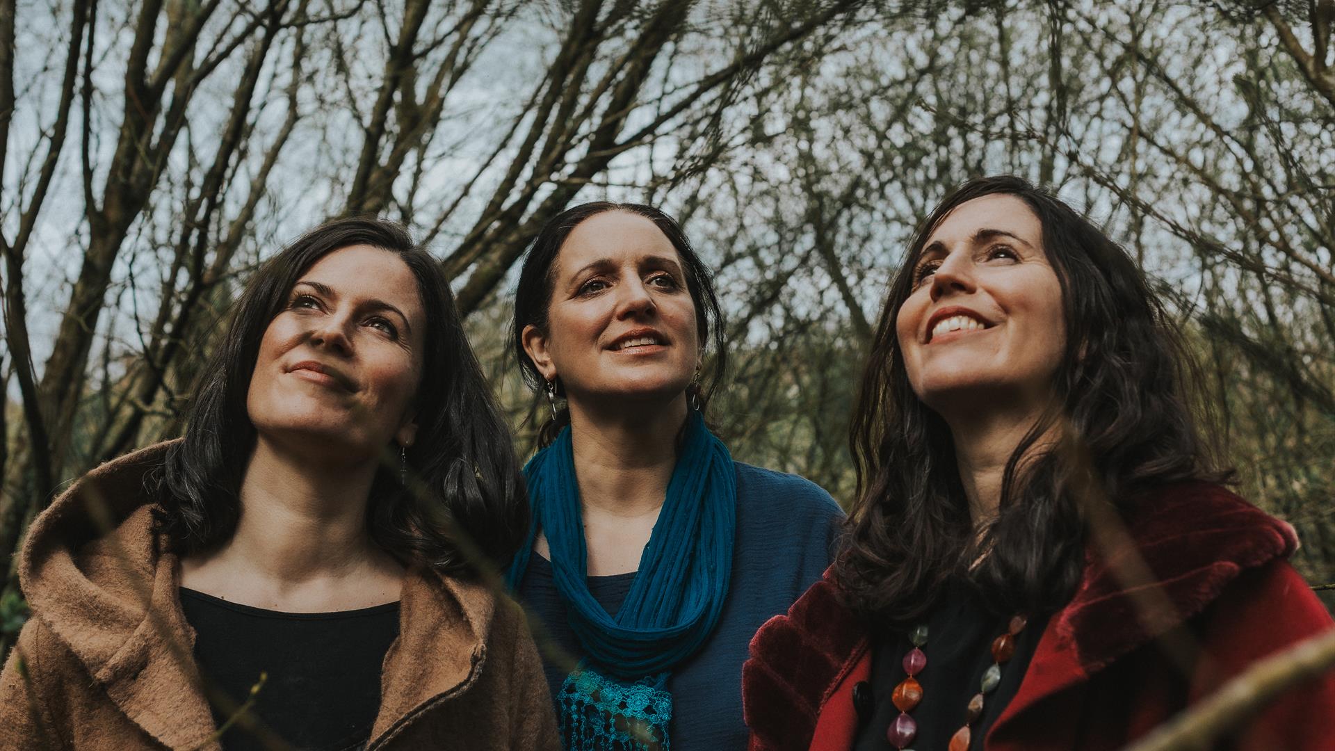 Image shows the Henry Girls, 3 women standing outside all smiling and looking upwards.