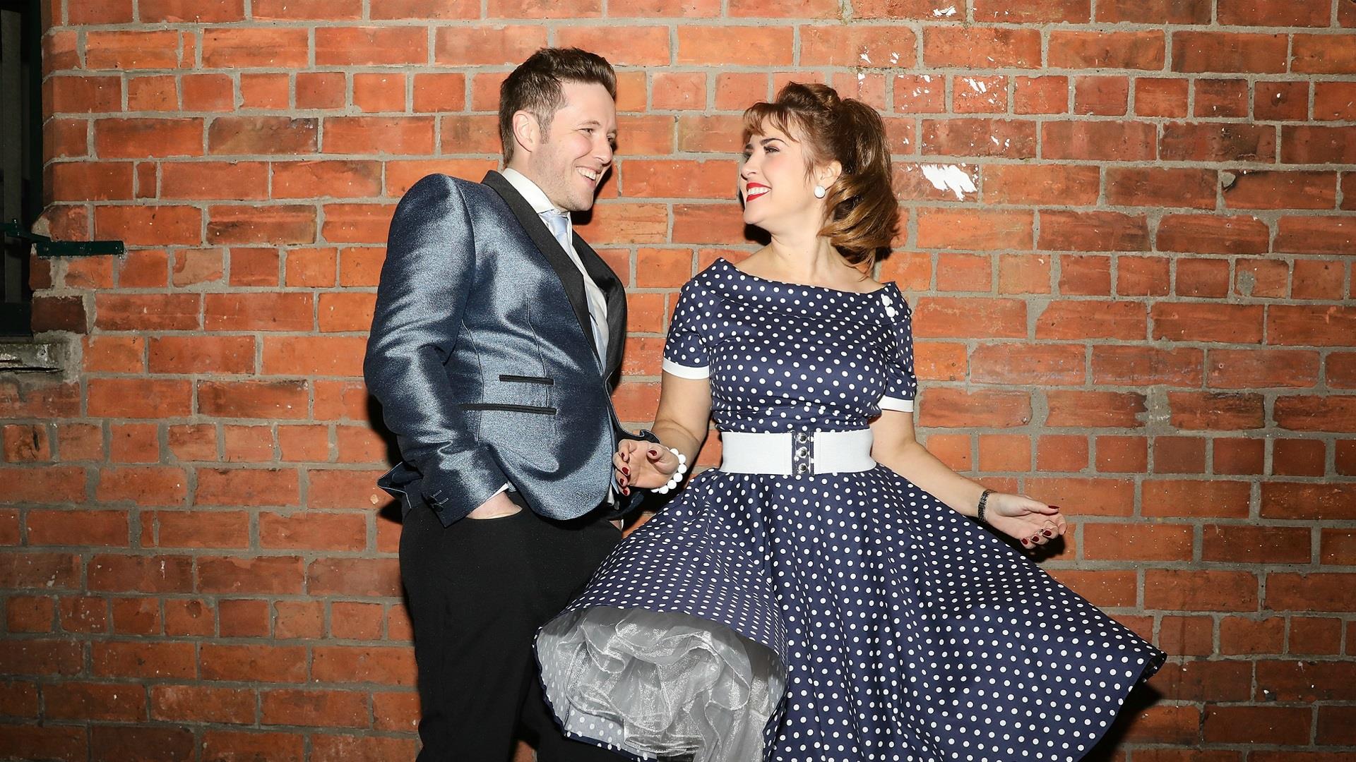 Picture of a man and a woman smiling at each other leading up against a wall. She is wearing a navy and white polka dot dress with a wide white belt,