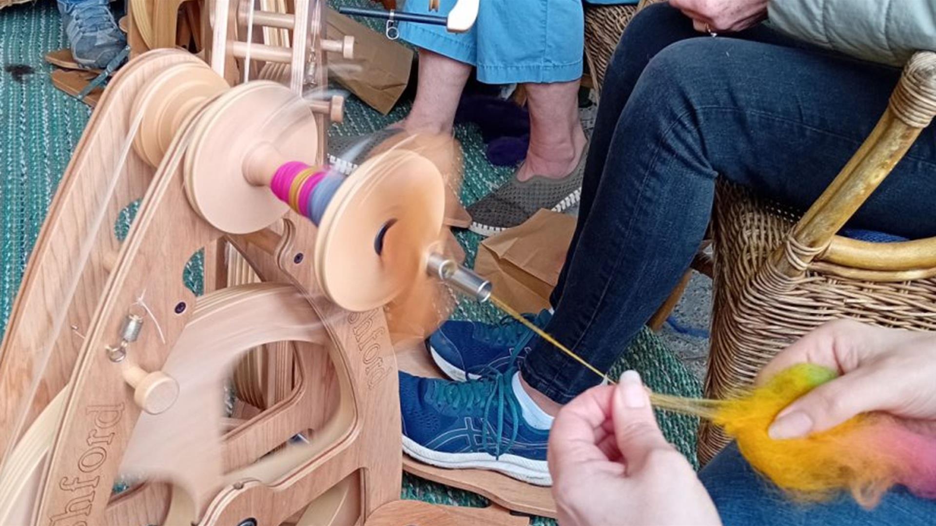 a group of people are seated and in the foreground, a person uses their hands to feed wool through a traditional wool spinning wheel a group of people are seated and in the foreground, a person uses their hands to feed wool through a traditional wool spinning wheel