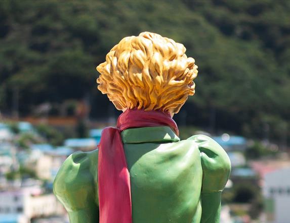 Image shows the back of a ceramic sculpture of a boy with a green jacket and blone hair and a red scarf.  Looking out over a blurry sunny city scape.
