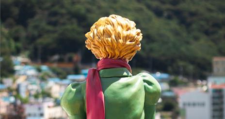 Image shows the back of a ceramic sculpture of a boy with a green jacket and blone hair and a red scarf.  Looking out over a blurry sunny city scape.