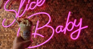 Moolicious milkshake topped with cream held up in front of a pink neon sign that says 'slice baby'.