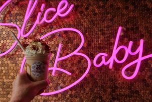 Moolicious milkshake topped with cream held up in front of a pink neon sign that says 'slice baby'.