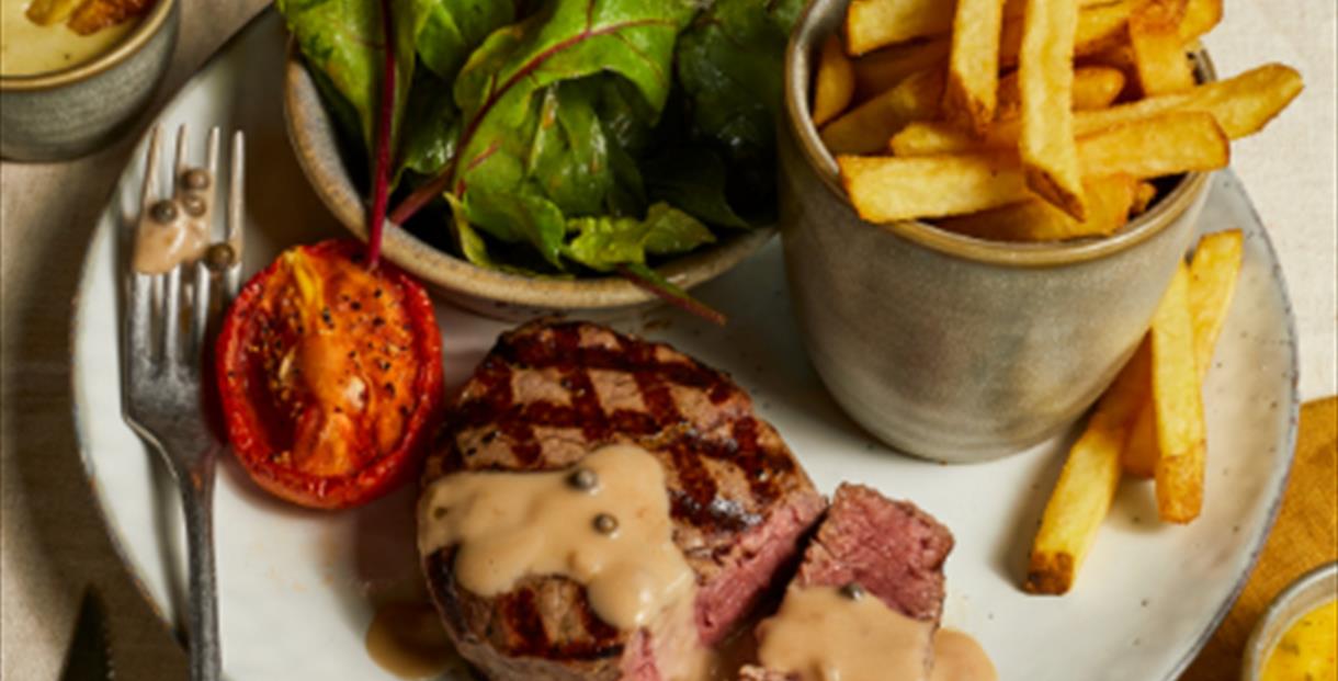 A seared steak with peppercorn sauce accompanied with grilled tomato, chips and green leaves