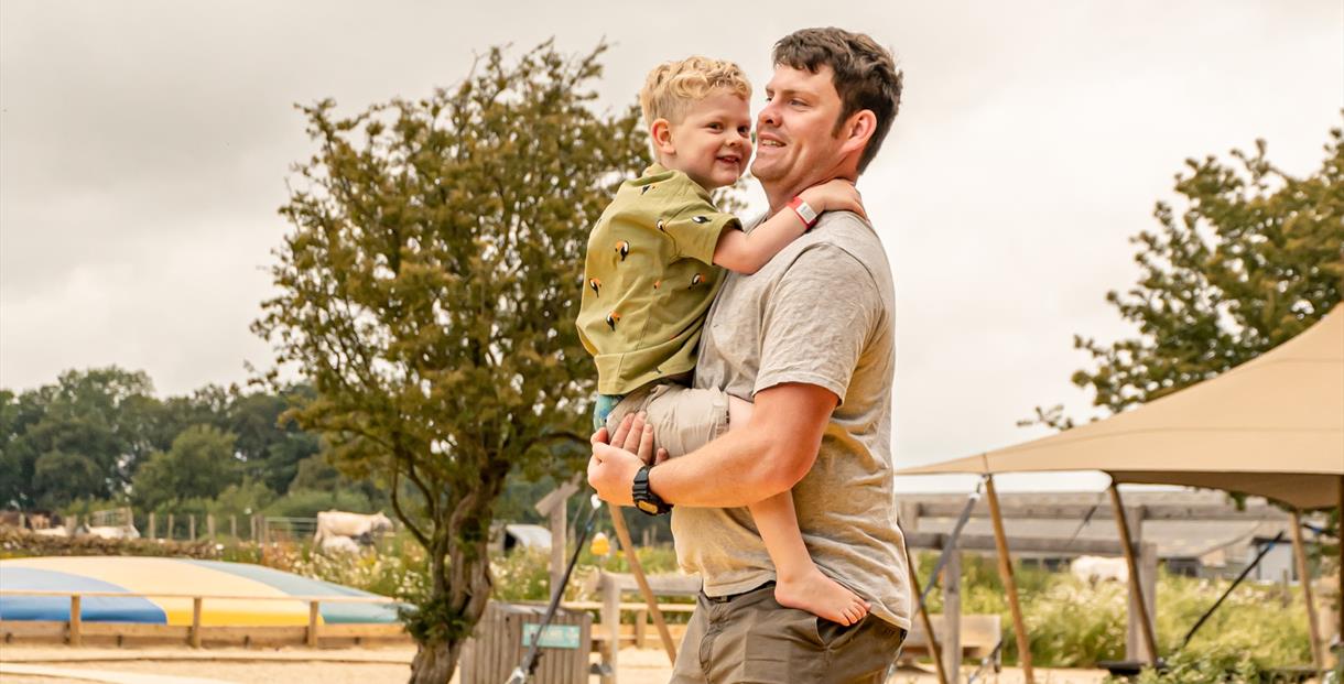 A man stands on the inflatable bouncy pillow at Cotswold Farm Park holding his son in his arms. The young boy is holding on to his dads shoulders and