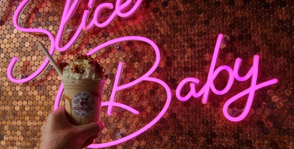 Moolicious milkshake topped with cream held up in front of a pink neon sign that says 'slice baby'.