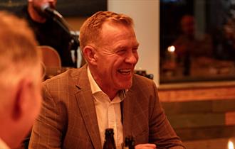 Adam Henson sits at a table in a dinner jacket smiling off to one side. A musician sits behind him with a microphone.