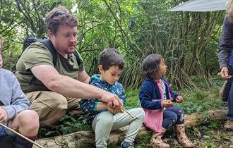 A dad and some children sitting on a log learning using peelers to sharpen sticks.