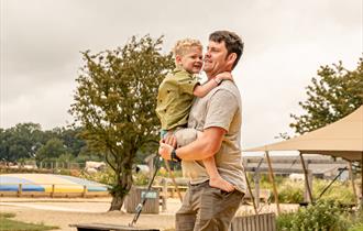 A man stands on the inflatable bouncy pillow at Cotswold Farm Park holding his son in his arms. The young boy is holding on to his dads shoulders and