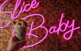 Moolicious milkshake topped with cream held up in front of a pink neon sign that says 'slice baby'.