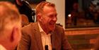 Adam Henson sits at a table in a dinner jacket smiling off to one side. A musician sits behind him with a microphone.