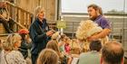 A male Cotswold Farm Park employee with long hair stands with a freshly shorn sheep's fleece under his arm. A crowd sits in front of him in a barn, wh