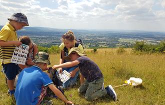 A family looks at the bugs they have found using a guide, beyond them there are far reaching views from the top of Cleeve Common over the low land bel