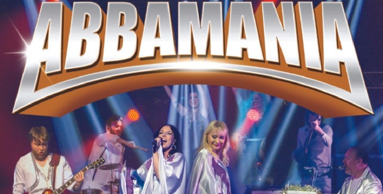 The female band members are centre stage singing while the other band members are to either side of them playing instruments, with the title Abbamania