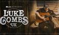 Luke Combs tribute performer sat in a pine cladded room playing an acoustic guitar, with an American flag hung on the wall behind them.