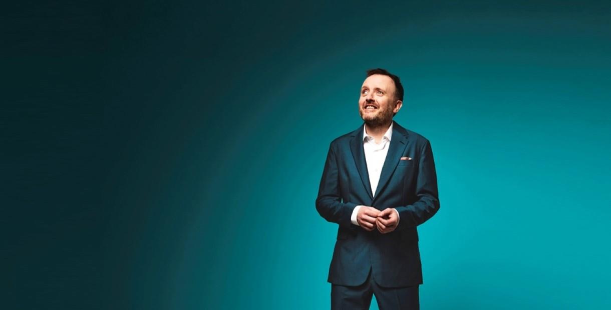 Comedian Chris McCausland wearing a suit in front of a teal background