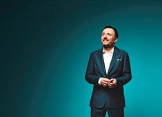 Comedian Chris McCausland wearing a suit in front of a teal background