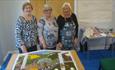 Three ladies stood behind a Well Dressing laid on a table