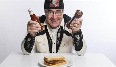 Danny Baker holding bottles of sauce over a sausage sandwich