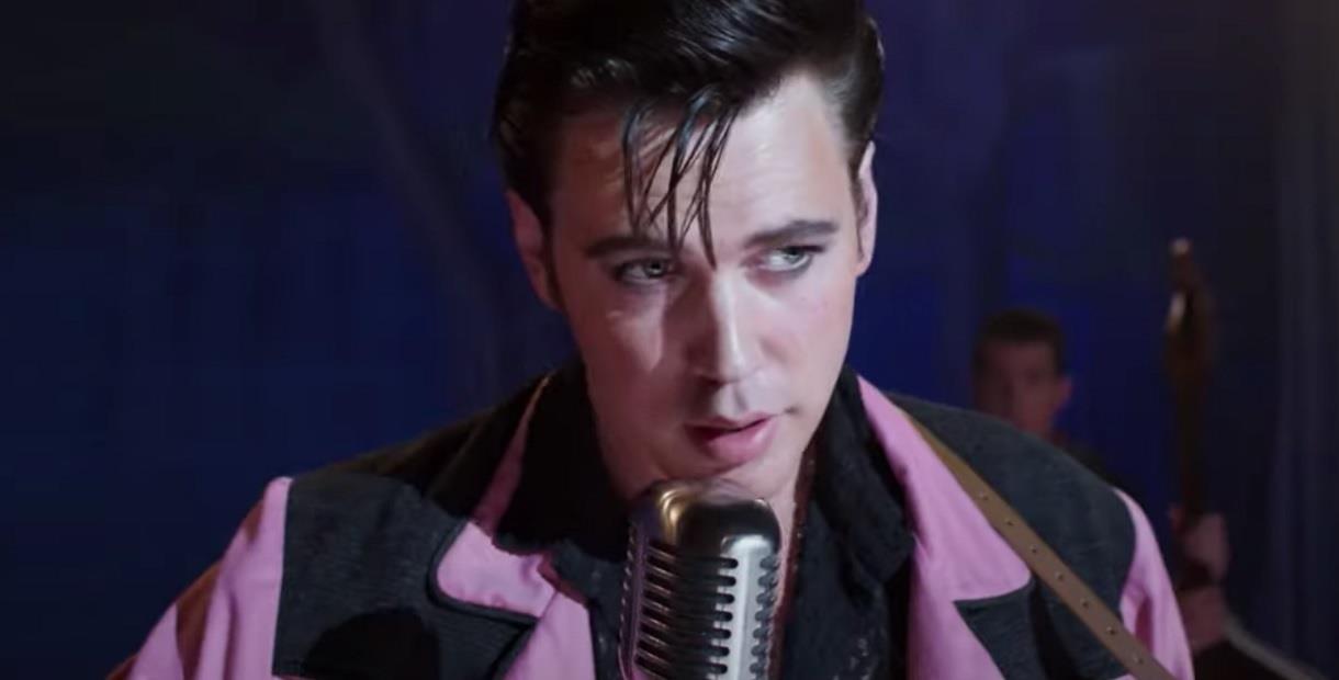 Austin Butler as Elvis in a pink and black suit, stands at the mic.