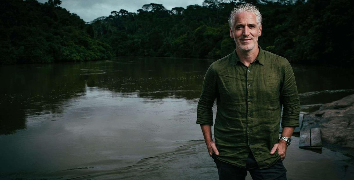 Gordon Buchanan stands on the shore of a lake, relaxed, hands in his pockets and smiling at the camera