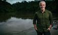 Gordon Buchanan stands on the shore of a lake, relaxed, hands in his pockets and smiling at the camera