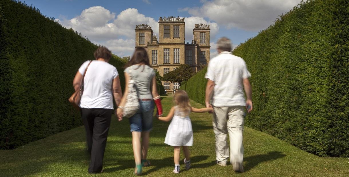 A family walk holding hands with a young girl between tall hedges towards Hardwick House