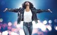 Naomi Ackie as Whitney Houston in jeans and a denim jacket looking up with her arms out