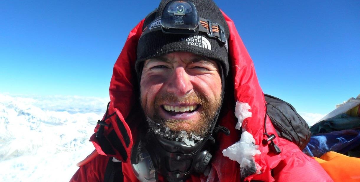 Adventurer James Ketchell smiling and looking very red faced, wearing a red hooded jacket, with mountains in the background