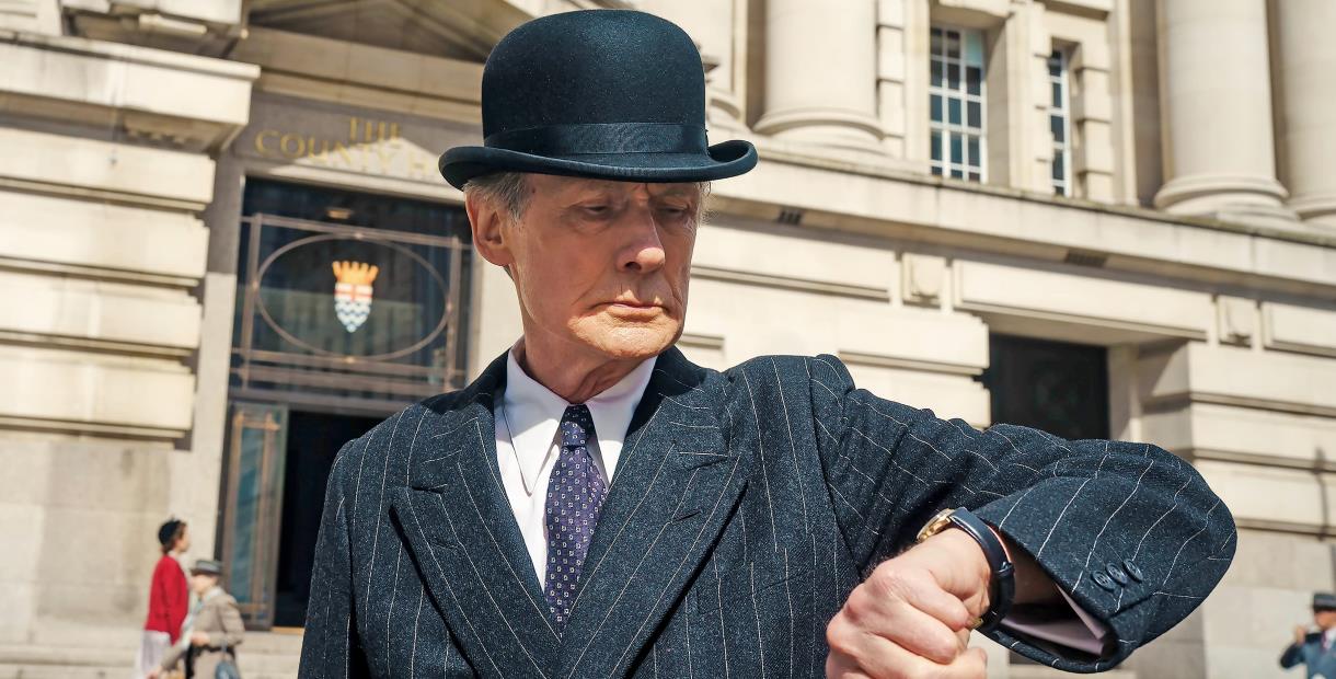 Bill Nighy wearing a pinstripe suit and bowler hat looking at his watch