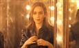 Emilia Clarke buttons her shirt while looking in the dressing room mirror with lights all around it.