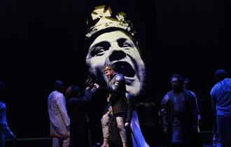 Actors in the foreground as the face of Richard the third looms large on a screen behind.