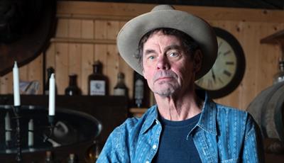 Rich Hall wearing a blue shirt and grey brimmed hat
