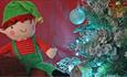 A close up of a Christmas elf toy next to a Christmas Tree.