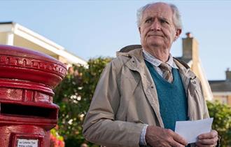 Jim Broadbent stood next to a red pillar post box, with a letter in his hand and a pensive look on his face.