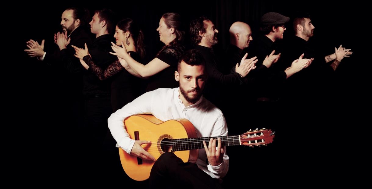Daniel Martinez is sat wearing a white shirt playing Spanish guitar while looking at the camera. The back ground is black with band members all wearin