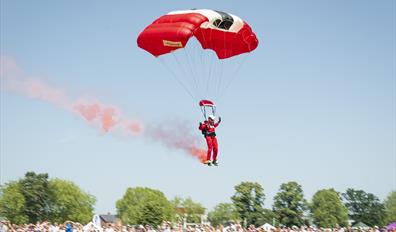 A parachutists witha  stream of red smoke lands in front of a crowd