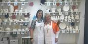 Two ladies in Jeans Craft Room with shelves of unpainted ceramics behind them.