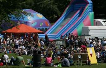 Large crowd of people enjoying the sunshine, with a bouncy castle and inflatables behind them.