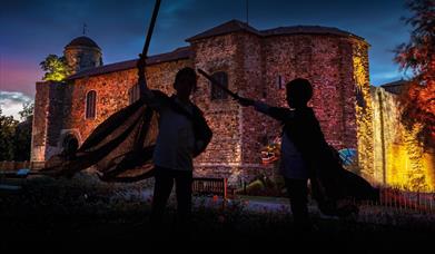 A silhouette of two children with toy swords in front of Colchester Castle
