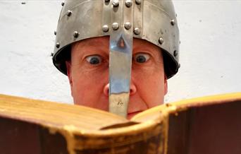 A man wearing a knight's helmet reads from a book