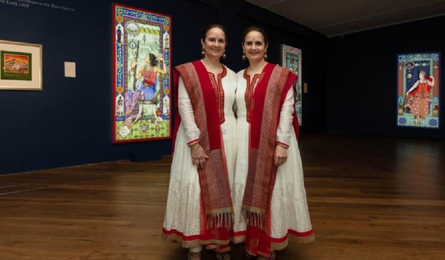 Two women in traditional Indian costume stand next to each other giving a somewhat symmetrical appearance. Illuminated artworks can be seen in the bac