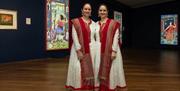 Two women in traditional Indian costume stand next to each other giving a somewhat symmetrical appearance. Illuminated artworks can be seen in the bac