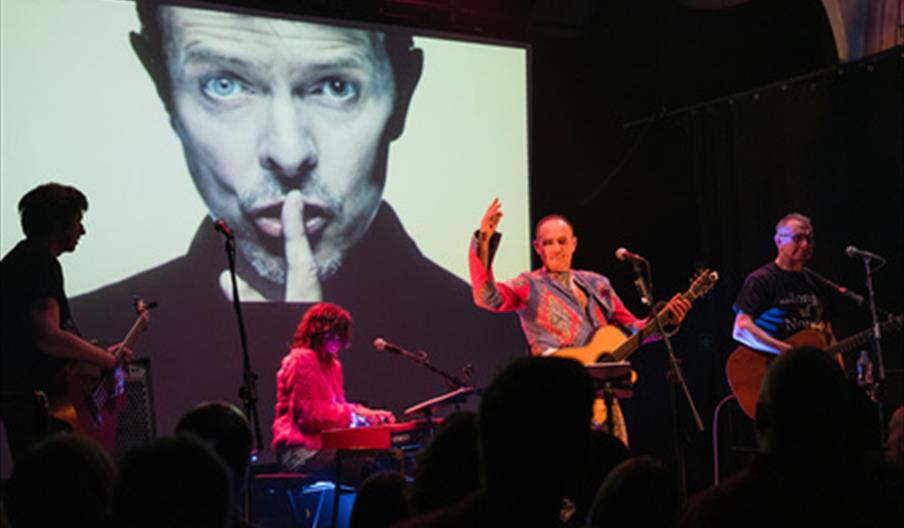 A live band performing in front of an image of David Bowie