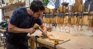 A person at a workbench using a hammer and chisel to carve a leaf design in wood