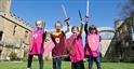 A groups of children dressed as knights raise their wooden swords in the air outside Sudeley Castle