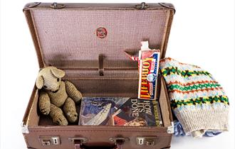 Evacuee's leather suitcase against a white background. The suitcase is open showing its contents: clothing, books, toiletries and a teddy bear